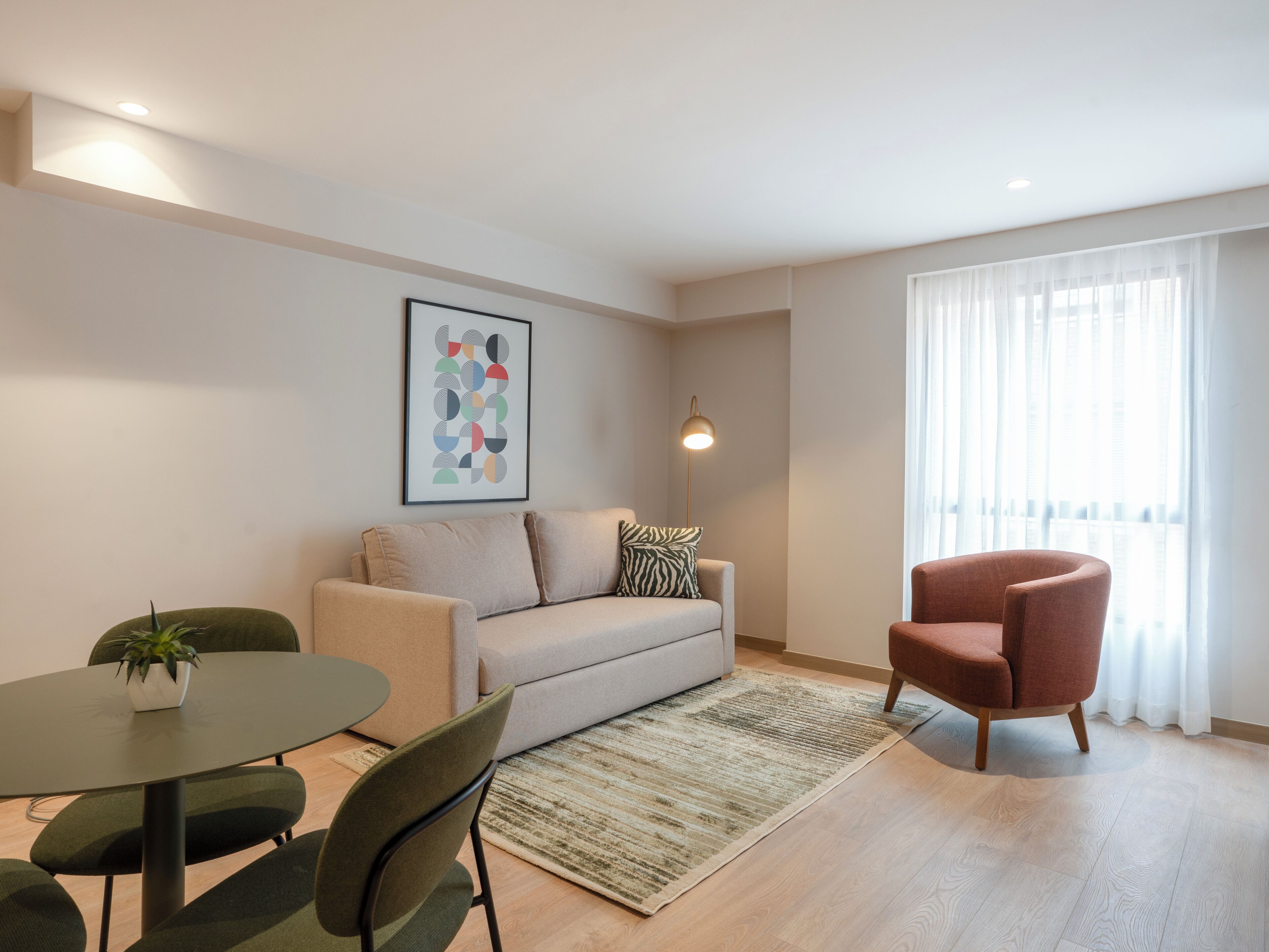 Una sala de estar moderna con sofá, un sillón rojo, una mesa redonda con sillas, una lámpara de pie y un ventanal con cortinas translúcidas.