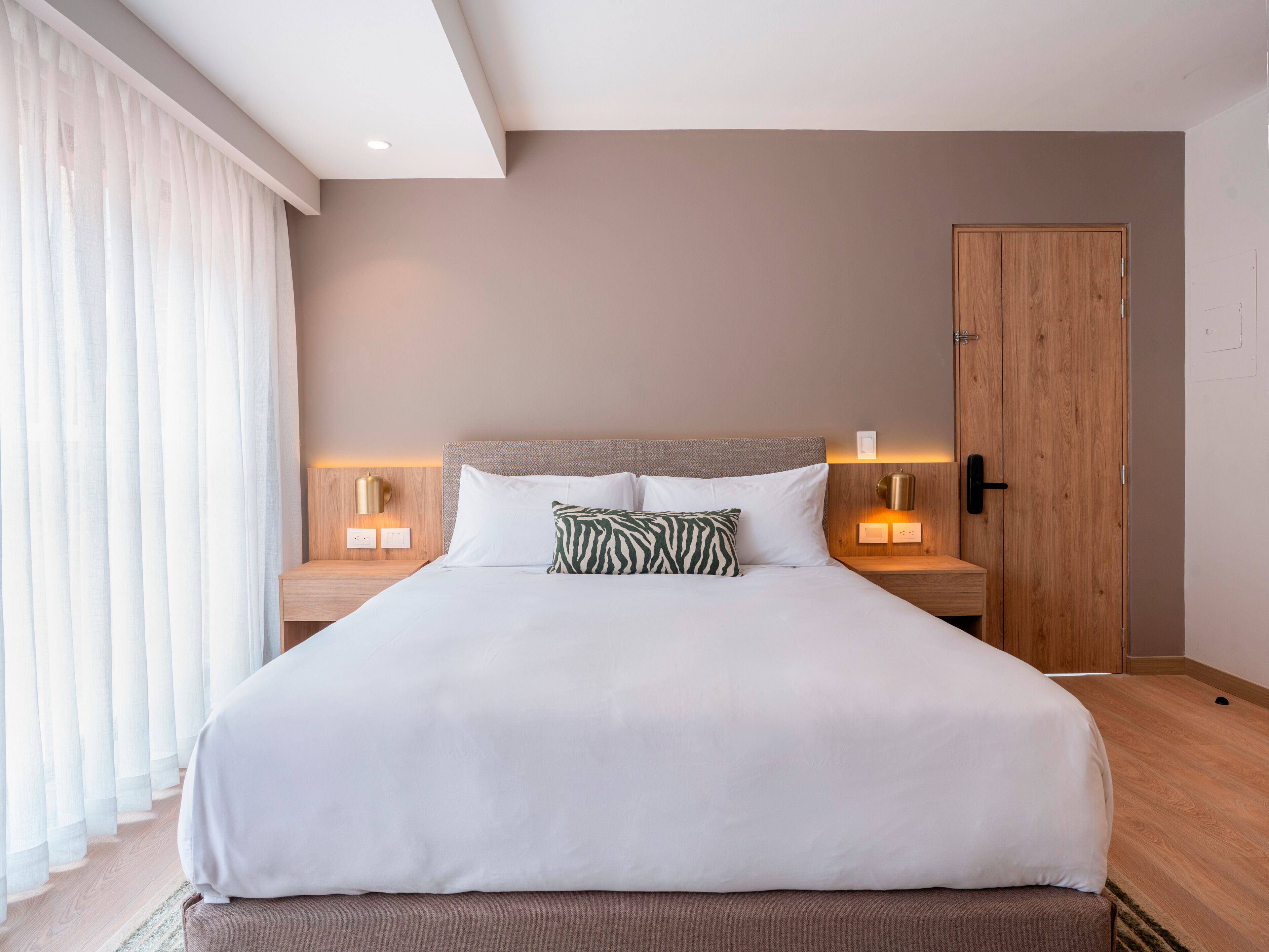 Una cama bien hecha con sábanas blancas y un cojín estampado con diseño de cebra, dos lámparas de noche, una puerta de madera y una ventana con cortinas de gasa.