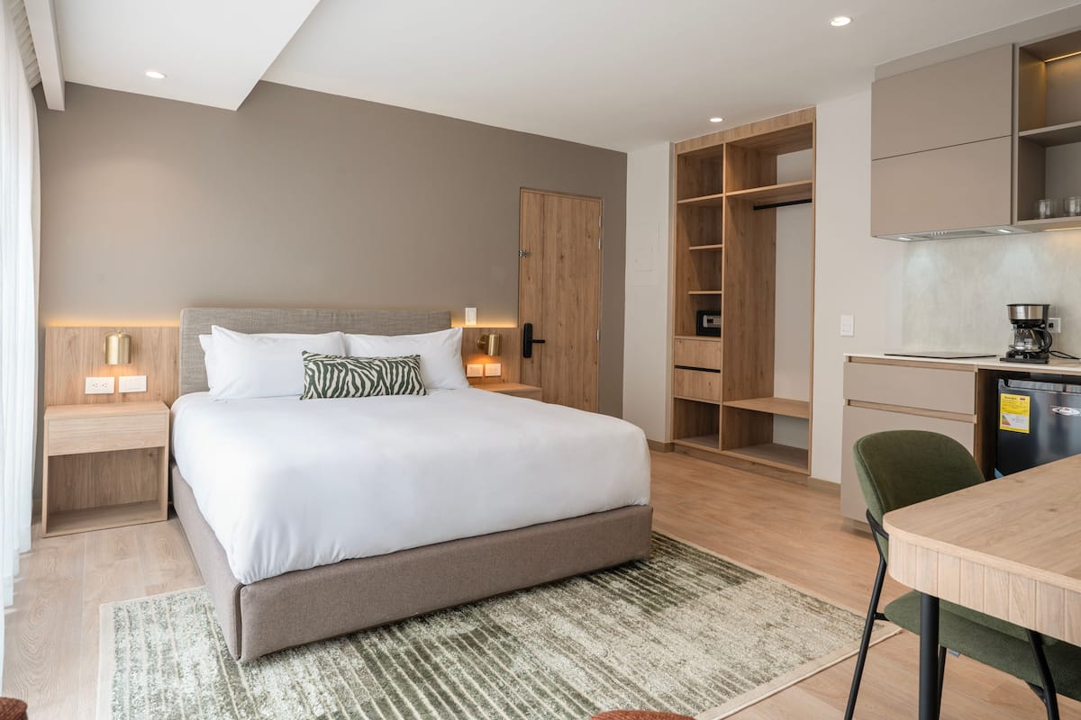 Una habitación de hotel moderna con una cama grande, lámparas de noche, un armario de madera, una kitchenette y un comedor.