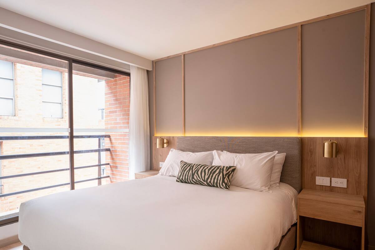 Una habitación de hotel moderna con una cama grande, una mesita de noche de madera, una lámpara de pared y vistas a un edificio de ladrillo a través de un ventanal amplio.