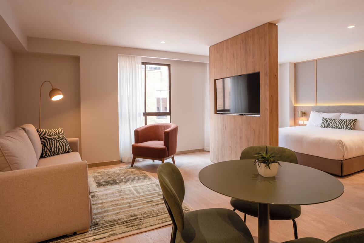 Una habitación de hotel moderna con sofá, sillón, mesa redonda y cama con televisor montado en la pared.