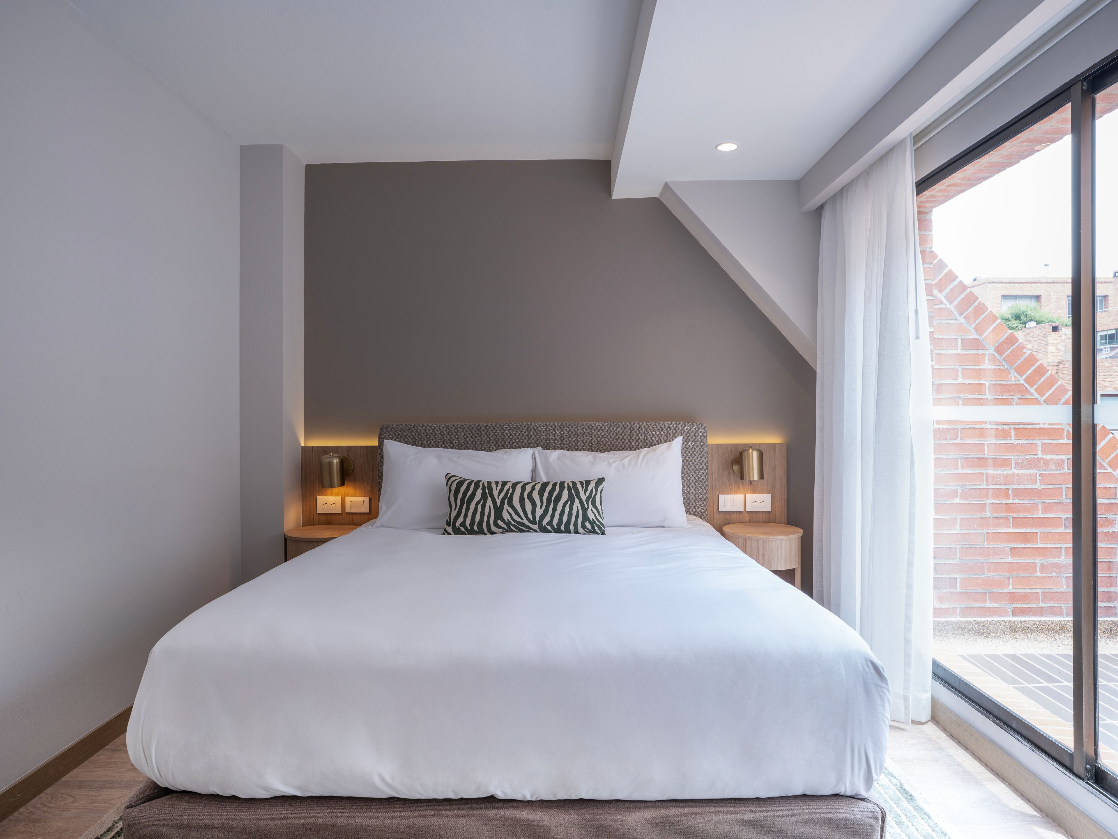 Una habitación de hotel moderna con una cama grande, dos mesitas de noche con lámparas, un techo inclinado y vista a un edificio de ladrillo a través de un ventanal amplio.