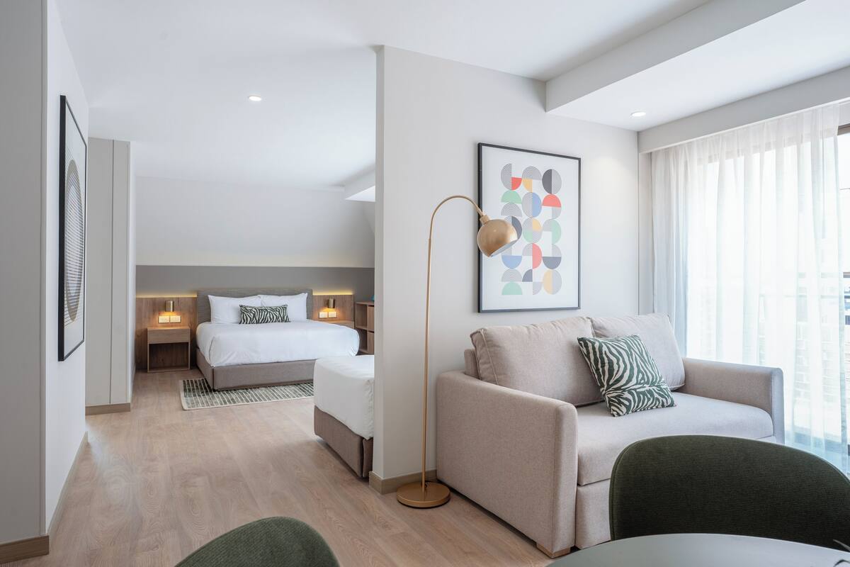 Una habitación de hotel moderna con cama, sofá, mesa de comedor y un amplio ventanal con cortinas.