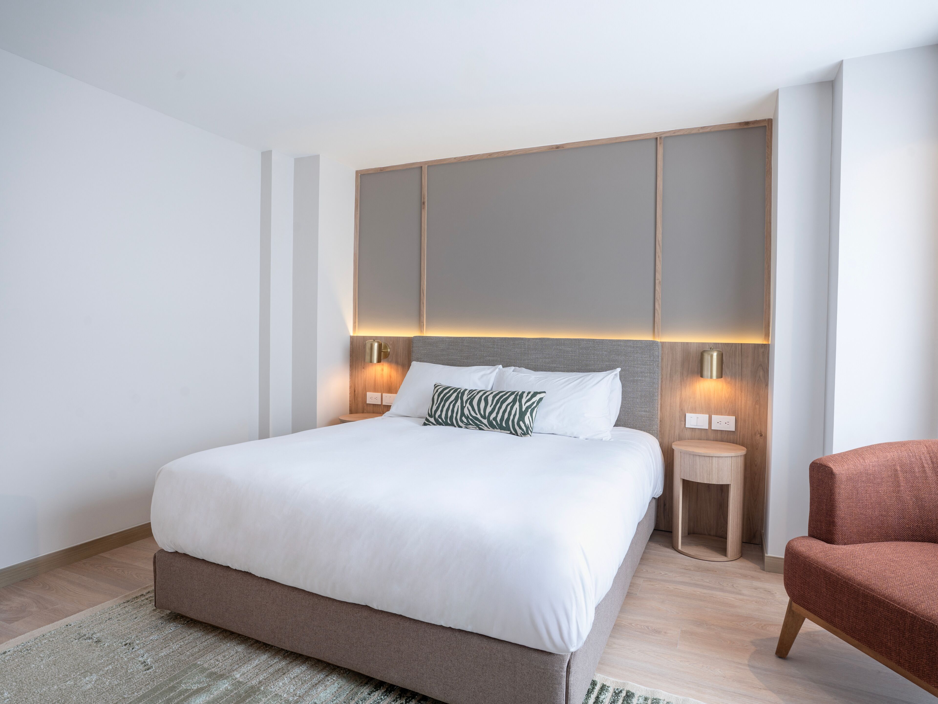 Una habitación de hotel moderna con una cama grande, dos mesitas de noche, una silla roja y un cabecero de madera.