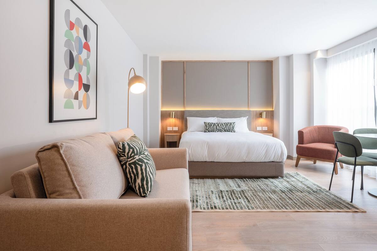 Una habitación de hotel moderna con una cama, un sofá, una silla, una alfombra y una obra de arte en la pared.