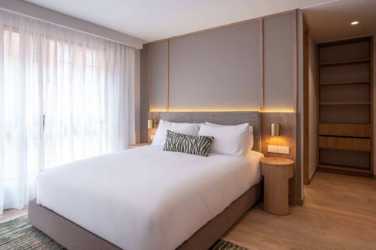 Una habitación de hotel moderna con una cama grande, lámparas de noche, un cabecero de madera y una estantería empotrada.