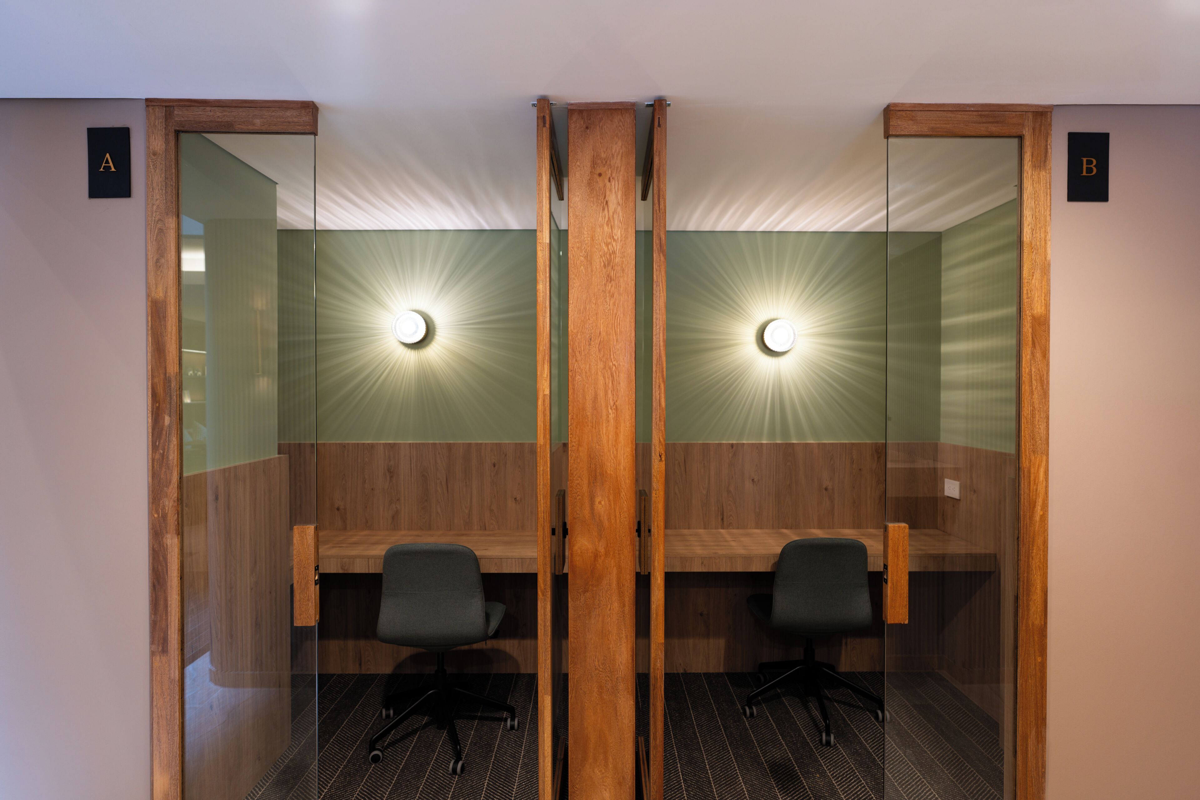 A mirrored room with two desks, each with a chair, and illuminated by wall-mounted lights.