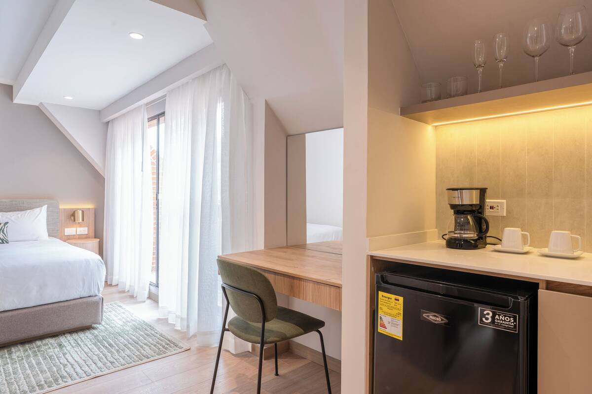 Una habitación de hotel moderna con una cama, un escritorio con cafetera, un refrigerador y una silla.