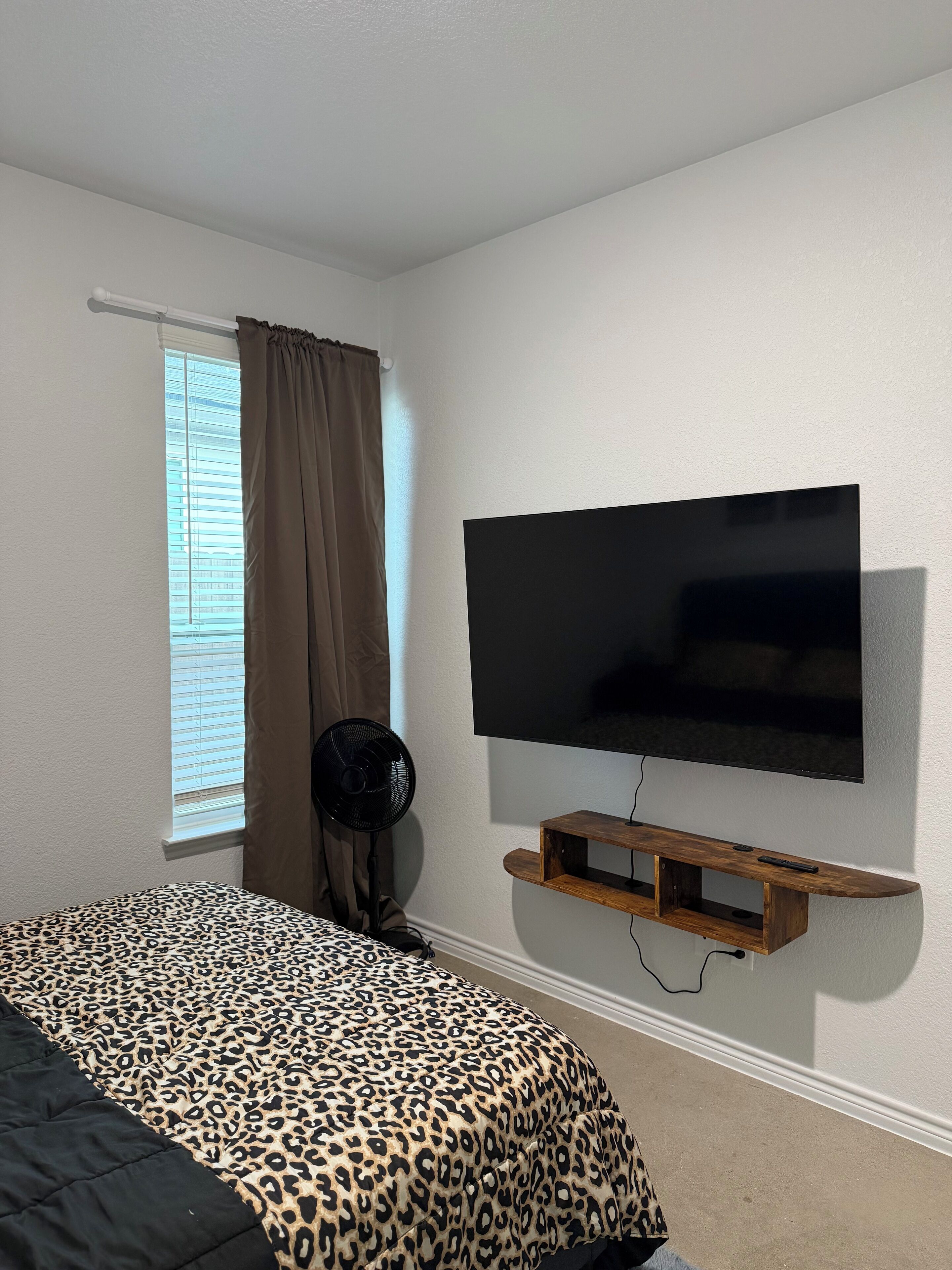 Une chambre à coucher avec un lit, un téléviseur fixé au mur, une fenêtre avec des rideaux et un ventilateur.