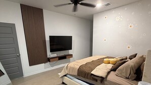 Un dormitorio con una cama, ventilador de techo, televisor, mesita de noche de madera y una silla.