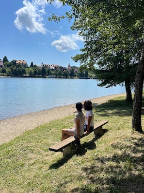 Deux personnes sont assises sur un banc au bord d’un lac, avec une ville en arrière-plan.
