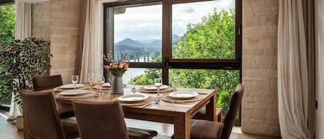 A dining area with a large table set for six, a potted plant, and a view of greenery and mountains through large windows.