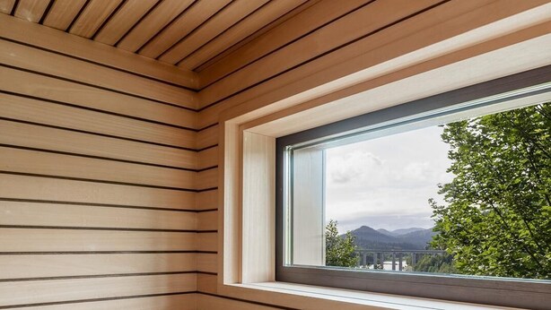 Una sala de sauna con listones de madera, una ventana con vistas a zonas verdes y montañas, y un cubo de madera.