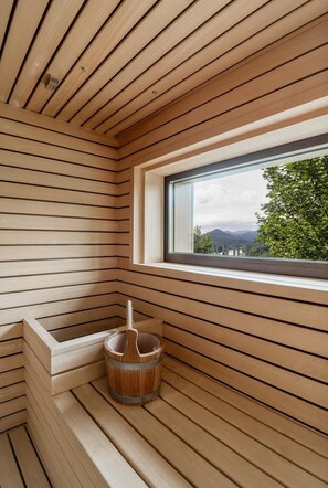 A sauna room with wooden slats, a window with a view of greenery and mountains, and a wooden bucket. - Villa Fužine Lake Luxury Retreat & Spa (Fužine)