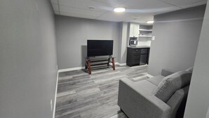 Ein modernes Wohnzimmer mit einem Flachbildfernseher auf einem Holzboden, einem grauen Sofa und einer Küchenzeile mit weißen Schränken und einer Mikrowelle.