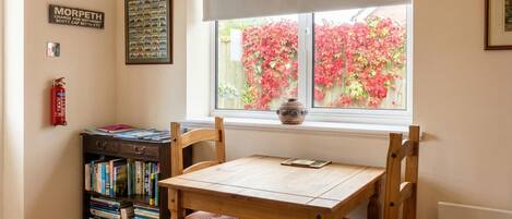 A dining area with a table and chairs, a bookshelf, a window with a view of a garden, and a fire extinguisher.