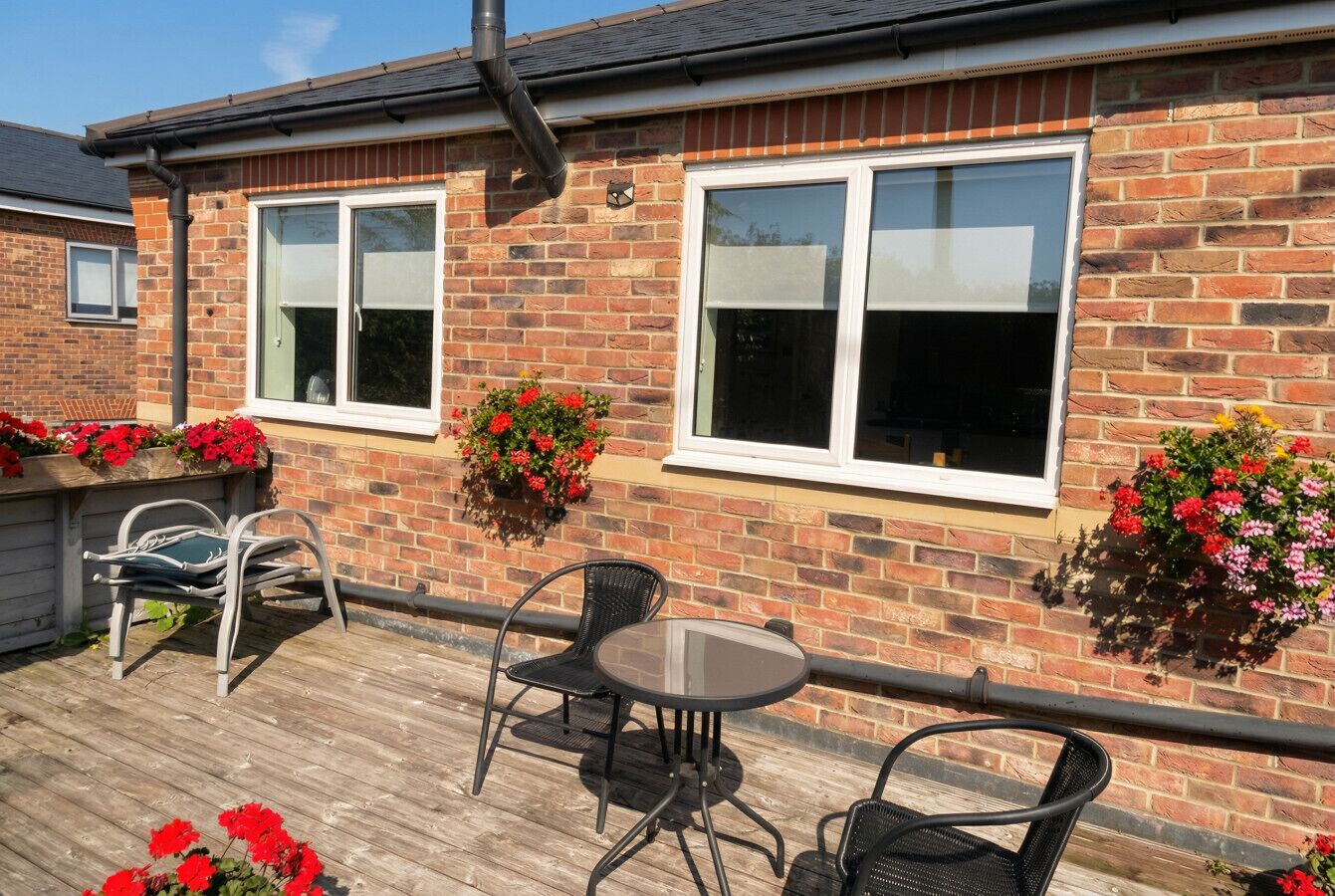 A brick house with two windows, a balcony with a table and chairs, and flower boxes on the wall.