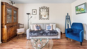 Ein Wohnzimmer mit grauem Sofa, blauem Sessel, Glastisch, Holzschrank und einem gerahmten Kunstwerk an der Wand.