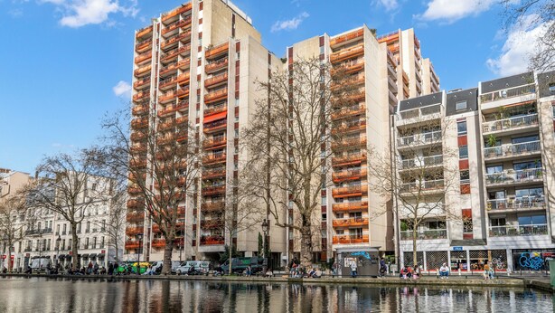 Ein Hochhaus mit mehreren Balkonen, ein Fluss im Vordergrund und ein klarer, blauer Himmel.