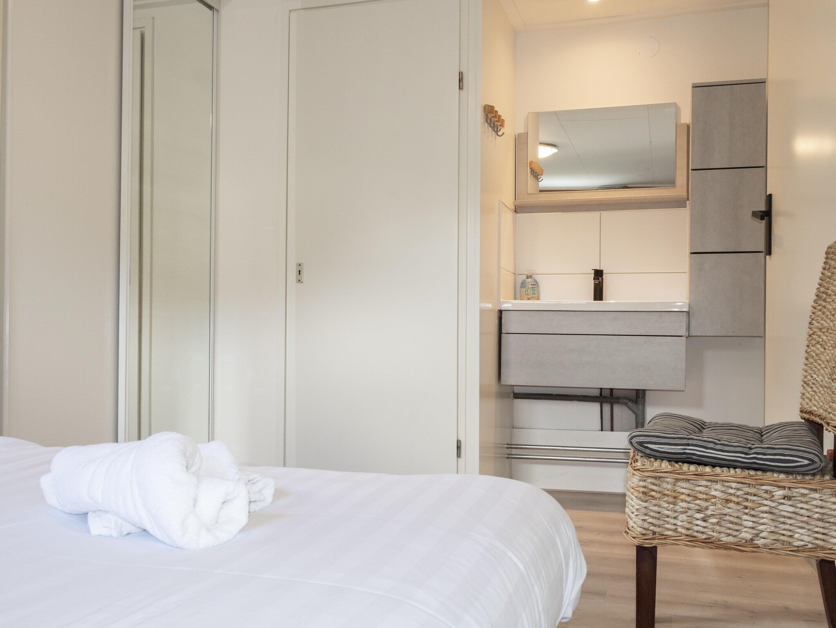Un dormitorio con cama, una silla, un armario y un baño con espejo y muebles.