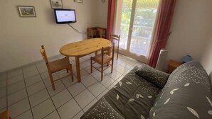 Ein kleiner, ordentlicher Raum mit einem Holzessttisch, zwei Stühlen, einem Sofa, einem an der Wand montierten Fernseher und einem Fenster mit Vorhängen.