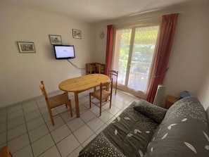 A small, tidy room with a wooden dining table, two chairs, a sofa, a television on the wall, and a window with curtains. - Air-conditioned studio with balcony in St-Raphaël (Saint-Raphaël)