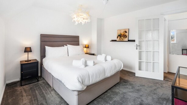 Habitación de hotel con una cama grande, lámparas de noche, un cuadro en la pared, una puerta con paneles de vidrio esmerilado y una pequeña mesa con una silla.