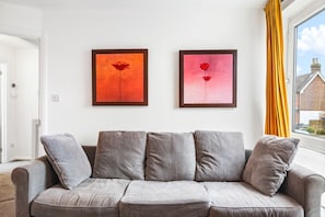 Un sofá gris con cojines, dos cuadros enmarcados en la pared y una ventana con cortinas amarillas.