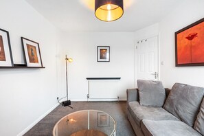 Una sala de estar con sofá, mesa de centro de vidrio, lámpara de pie, cuadros enmarcados en la pared, radiador y una puerta.