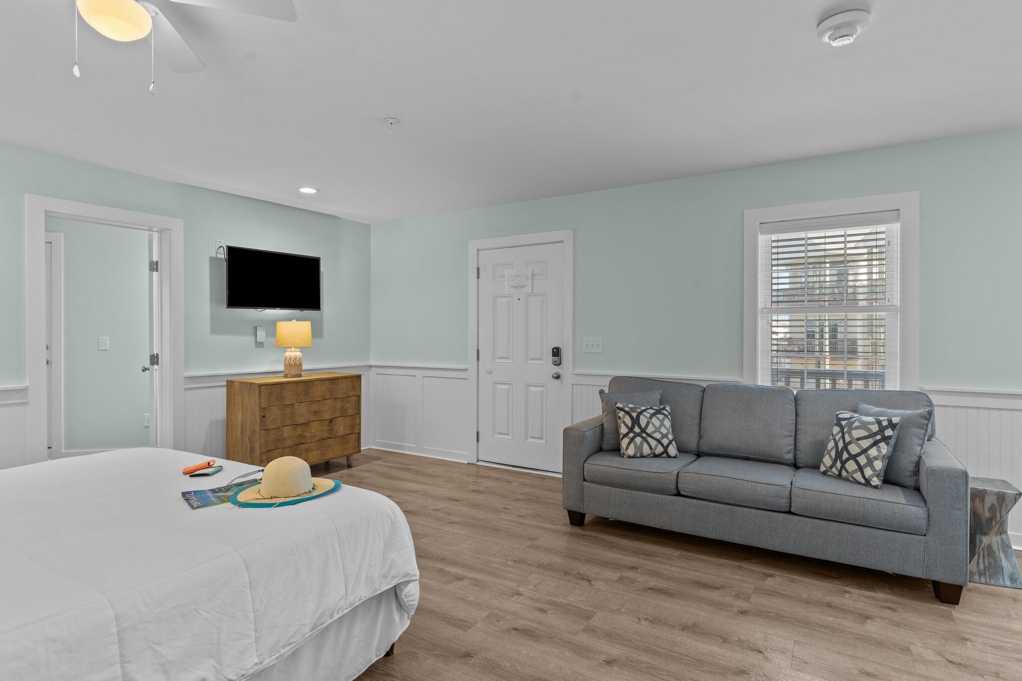 A bedroom with a bed, a gray sofa, a wooden nightstand, a TV mounted on the wall, and a window with blinds.