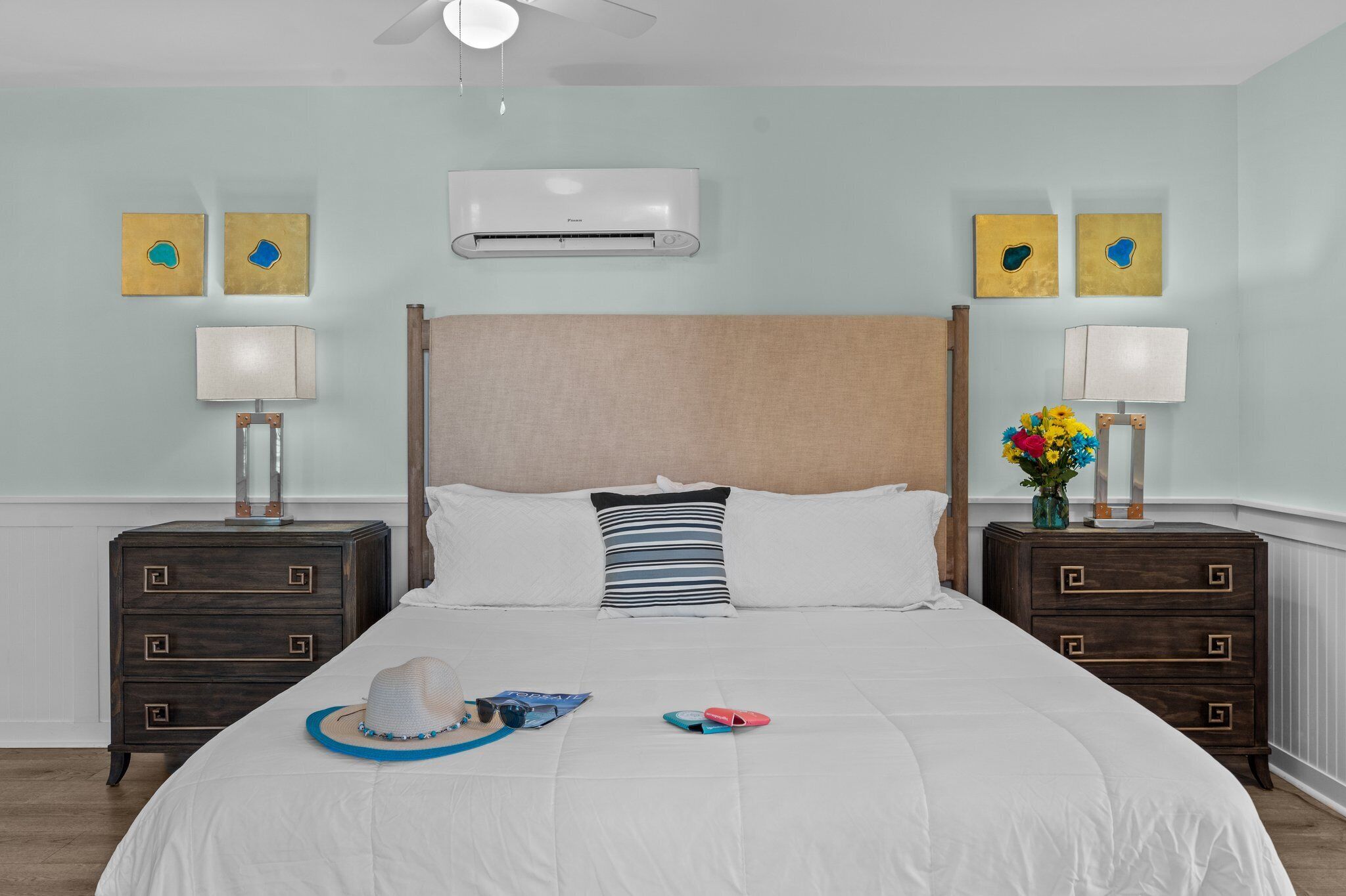 A neatly arranged bedroom with a large bed, two bedside tables with lamps, an air conditioning unit, and wall art.