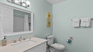 A bathroom with a white sink, two faucets, a mirror, a toilet, and two wall-mounted light fixtures. - Loggerhead Inn & Suites - King Suite in hotel in Surf City, North Carolina (Surf City)