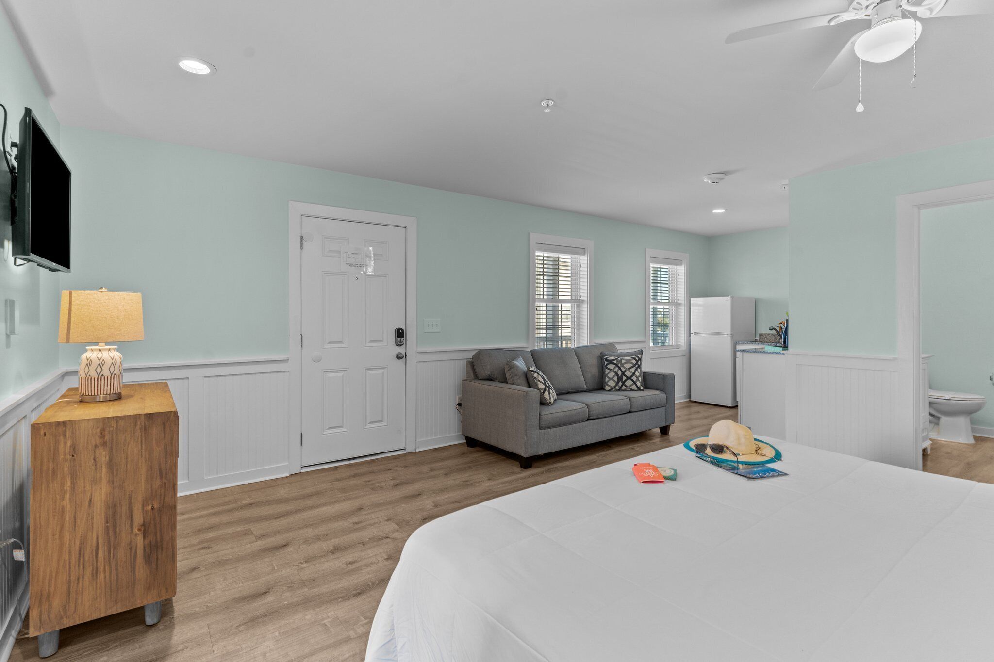 A bedroom with a bed, a wooden nightstand, a gray sofa, a television, and a ceiling fan.