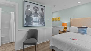 A hotel room with a bed, a chair, a nightstand with a lamp, a framed artwork on the wall, and a door. - Loggerhead Inn & Suites - King Premium Suite in hotel in Surf City, North Carolina (Surf City)