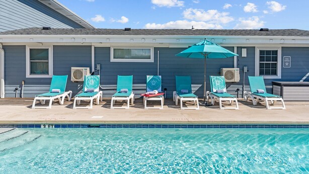 A poolside area with lounge chairs and an umbrella, a clear sky, and a building with a blue exterior. - Loggerhead Inn & Suites - King Premium Suite in hotel in Surf City, North Carolina (Surf City)