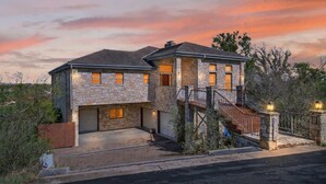 A two-story stone house with a garage, a covered driveway, and a wooden staircase leading to the entrance. - New! Luxury home with Views & Hot Tub, Game Room! (Horseshoe Bay)