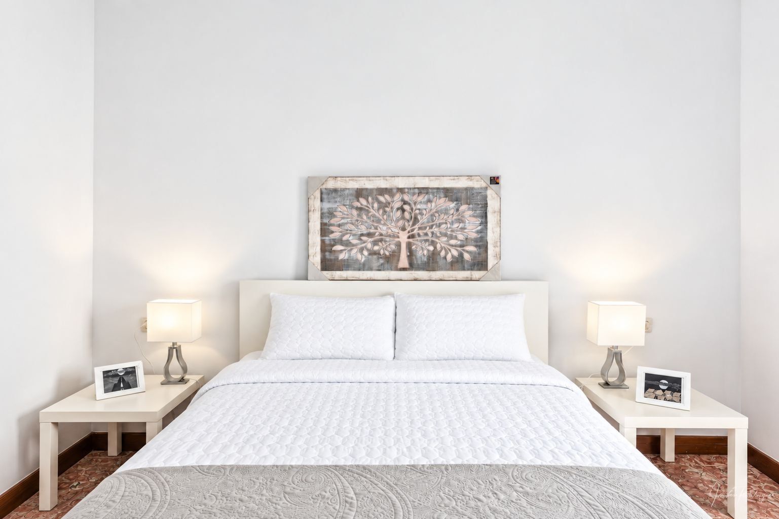 Una cama bien hecha con colcha blanca y dos almohadas, flanqueada por dos mesitas de noche con lámparas y fotografías enmarcadas.