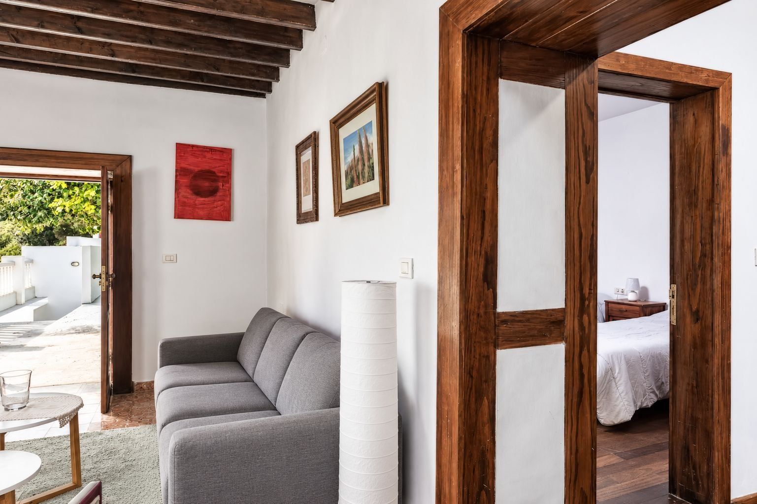 A room with a grey sofa, a white lamp, and a bed with a white cover. There are wooden doors and a white wall with framed pictures.