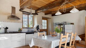 Una cocina con armarios blancos, una mesa de comedor con sillas, una lámpara colgante y una ventana con vistas a árboles.