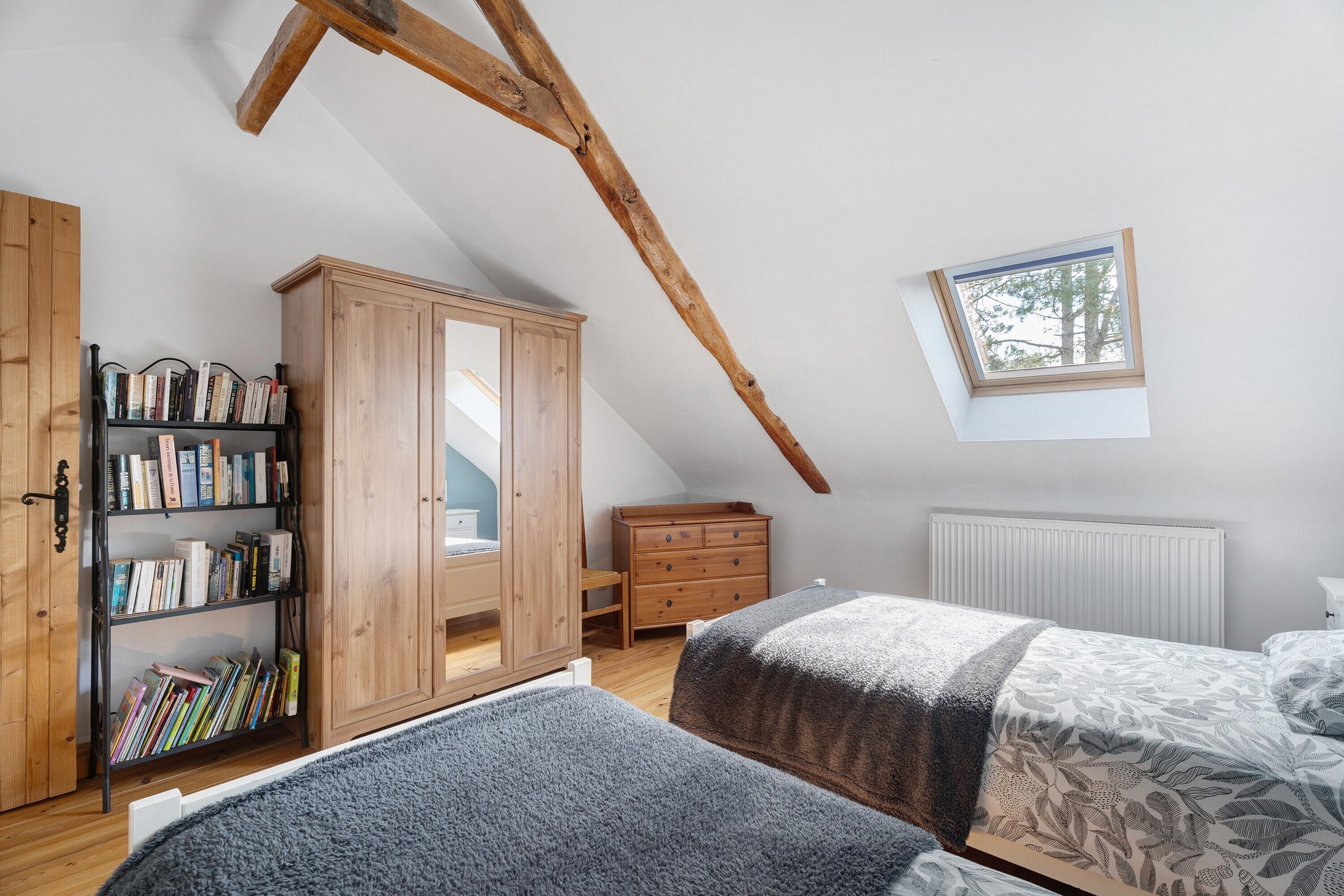 Een slaapkamer met een schuine kap, een houten garderobe, een boekenkast, een raam en een radiator.