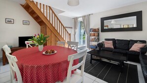 A dining area with a red polka dot tablecloth, white chairs, a wooden staircase, a black sofa, and a large mirror on the wall. - Cottage with Heated Indoor Pool (Saint-Thual)