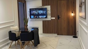 A modern living room with a flat-screen TV mounted on the wall, a glass coffee table, two chairs, a potted plant, and a wooden door. - 2BR City Center Medina With Terrace Rabat (Rabat)