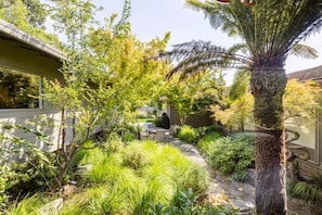 A lush garden with a variety of plants and a palm tree, a patio area with a table and chairs, and a building with a sloped roof.