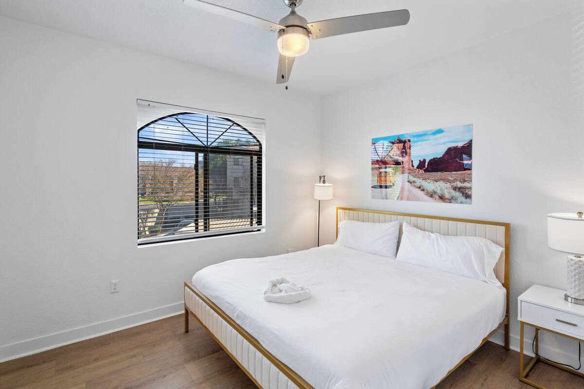 Une chambre à coucher comprenant un lit, un ventilateur de plafond, une fenêtre avec des stores, une table de chevet avec une lampe et un tableau accroché au mur.