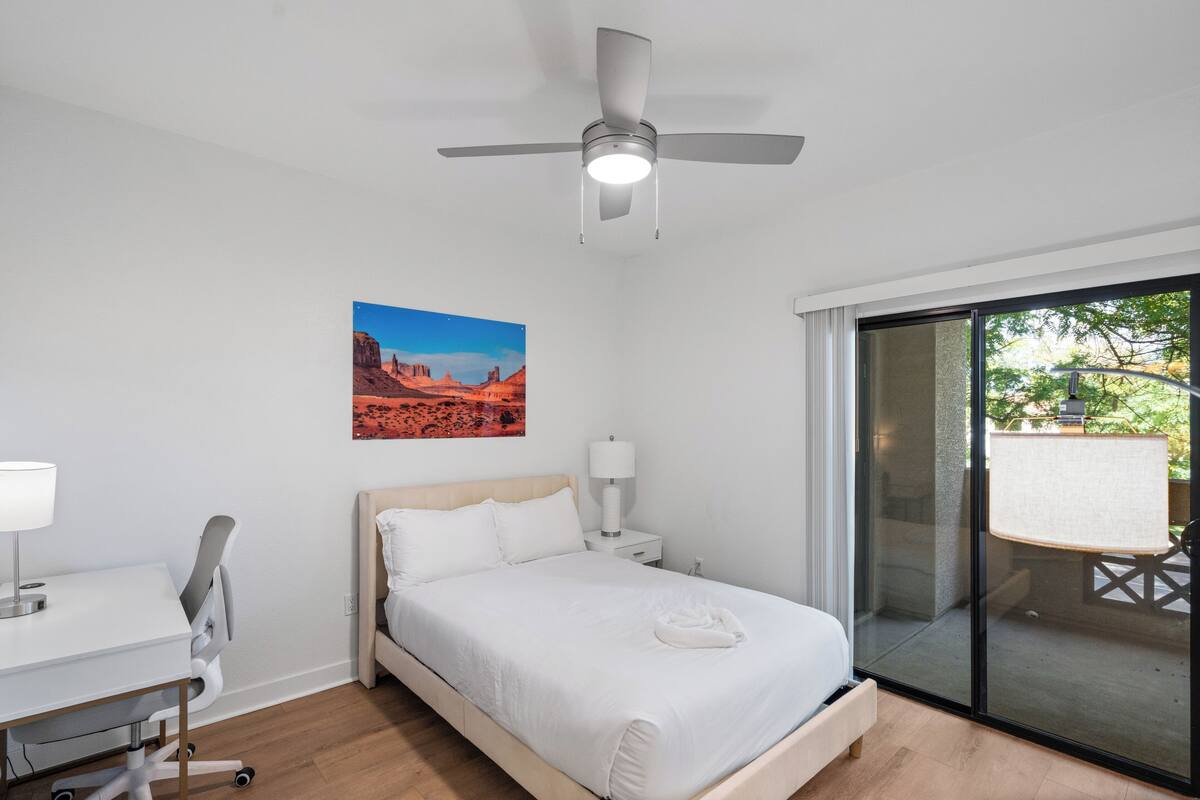 Une chambre à coucher comprenant un lit, un bureau, une chaise, un ventilateur de plafond, une table de chevet, une lampe, une fenêtre et un tableau.