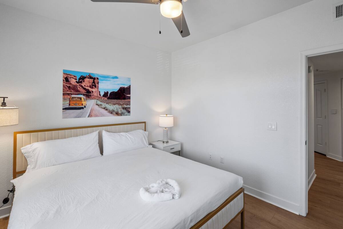 Une chambre d’hôtel comprenant un lit, une table de chevet, une lampe, un ventilateur de plafond et une porte donnant sur une autre pièce.