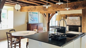 A rustic kitchen with exposed wooden beams, a central island with a black countertop, and hanging pendant lights. - Gîte du Alchimiste on the Château de Dompierre estate (Dompierre-les-Églises)