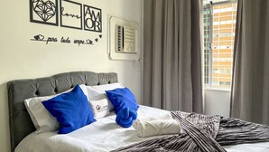 A neatly made bed with a grey headboard, a wall with geometric art and a heart-shaped phrase, and a window with curtains. - Rio Select - Copacabana  (Rio de Janeiro)