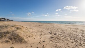 En bred sandstrand med fotspår och gles vegetation, som leder ner till ett lugnt hav under en klar himmel.