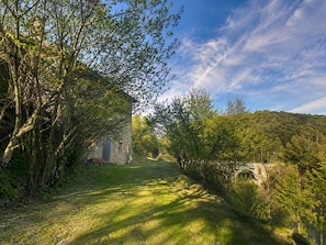 A rural pathway leading to a stone building with a rustic appearance, surrounded by lush greenery and a clear sky.